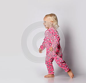 Laughing barefooted blonde baby kid girl in pink warm comfortable jumpsuit is running aside over grey wall background