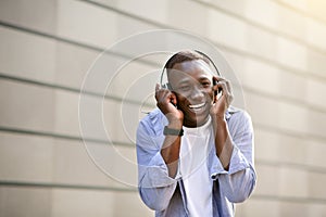 Laughing black guy listening to music in headphones near brick wall outside, blank space