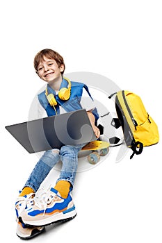 Laughing blonde boy sitting on yellow skateboard and holding notebook on white background. Back to School.