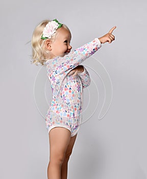 Laughing blonde kid baby girl in jumpsuit one-piece with floral pattern stands side to camera pointing at upper corner