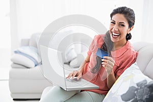 Laughing brunette sitting on her sofa using laptop to shop online