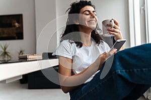 Laughing brunette woman using mobile phone while drinking coffee