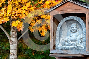 The Laughing Buddha, Budai, with his cloth sack stone sculpture with a yellow maple tree in the background