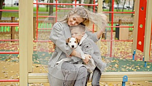 Laughing Caucasian mother and son hugging cute dog sitting on bench on playground. Portrait of happy cheerful family