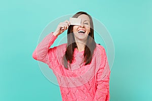 Laughing cheerful young woman in knitted pink sweater covering eye with credit card in hand isolated on blue turquoise
