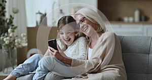 Laughing child enjoy pastime with granny watching videos on smartphone