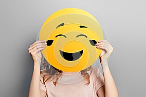 Laughing emoticon with tears of joy. Girl holds a yellow smiley with romantic love face emoticon  on a grey