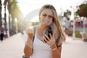 Laughing excited young woman watching her smartphone celebrating outdoor raising her fist up in exultation
