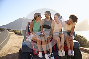 Laughing Female Friends Sitting On Hood Of Open Top Car On Road Trip