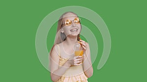 Laughing girl holding in hands orange juice glass on transparent green background. Happy girl teenager in yellow glasses