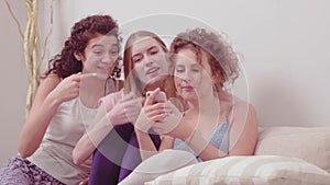 Laughing girls making selfies using smartphone or recording videos for social media in home clothes. Three girlfriends