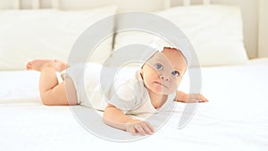 laughing happy little baby girl with blue eyes in a white bodysuit on her tummy on a bed in a bright bedroom on a cotton