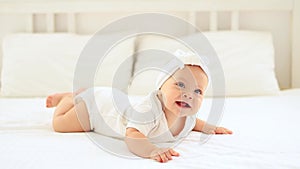 laughing happy little baby girl with blue eyes in a white bodysuit on her tummy on a bed in a bright bedroom on a cotton