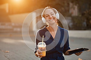 Laughing happy woman with drink and tablet. Pretty female listen music in headset. Coffee time and take break concept