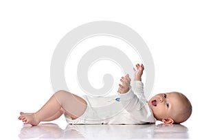 Laughing infant baby toddler in white bodysuit is lying on his back, holding hands up, waving, happy screaming on white