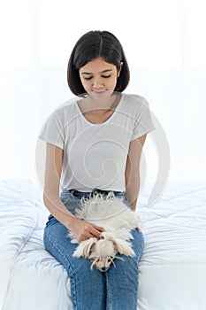 Laughing jocund young asian woman sitting on bed in bedroom with her maltese dog on knees