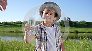 Laughing kid drinks mineral water from transparent glass at nature in summer