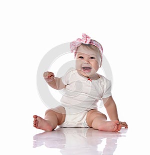 Laughing little baby infant girl in white body and hair bow decoration sits on floor looking at camera waving with hand