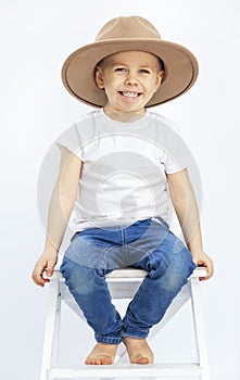 A laughing little boy in a white T-shirt, jeans and hat sits on a chair. Light background. Full height. Vertical