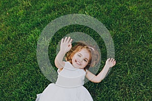Laughing little cute child baby girl in light dress lie on green grass lawn, showing palm on camera in park. Mother