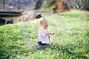 Laughing little girl with a ponytail on her head sits on her knees on a green lawn among white daisies