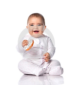 Laughing loudly infant baby toddler in white onepiece jumpsuit overall sits on the floor holding donut in hands waving