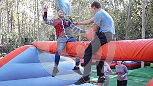 Laughing man sitting on inflatable log and fighting by pillows with his friend on adults bouncy playground