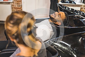 Laughing man using salon hair wash chair. Blurred head of another client in the foreground. Hairdresser concept.