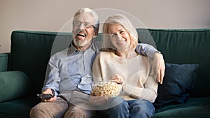 Laughing mature spouses sitting on couch watching comedy at home