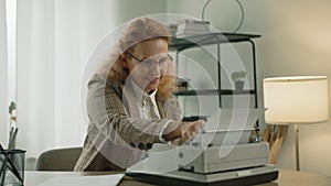 Laughing middle-aged blonde woman in glasses typing on typewriter at home