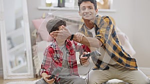 Laughing Middle Eastern teenager closing eyes of little brother playing video game using game controllers. Portrait of