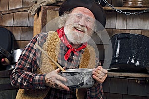 Laughing old cowboy cook holds a saucepan