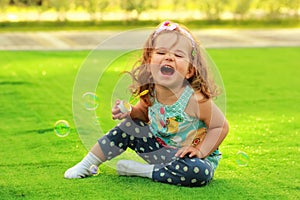 Laughing one year old girl learning to blow soap bubbles and sitting on the sunlit lawn