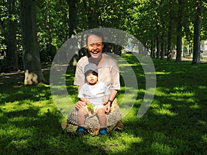 Laughing Papuan mom sitting with her toddler son on grass in alley of trees