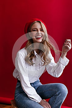 Laughing pretty woman in beret sitting on red background. Amazing girl in jeans and shirt smiling at camera