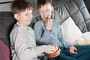 Laughing school aged boys sit on sofa, eat popcorn and watch comic TV show. Kids having fun and relaxing at home.