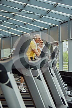 Laughing senior couple enjoys fun workout on treadmills in modern fitness center. Cardio training to fat burning.