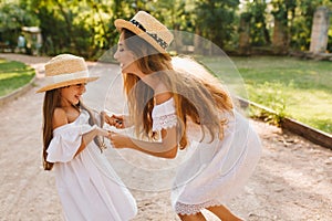 Laughing tanned woman bent over to smiling girl and dancing with her. Outdoor portrait of cute young mother and blissful