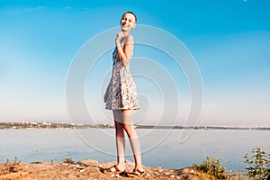 Laughing teenager girl in a short haired dress stands on a cliff over a wide river