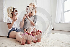 Laughing together. Pedicure and painted leg nails. Conception of skin care by using white mask and cucumbers on the face