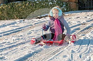 Laughing, vibrantly dressed young girl with blue ear muffs sledging down a slope