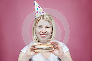 Laughing woman in home clothes and a festive cap is holding a hamburger. Lonely birthday in isolation period on the background of