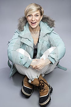 A laughing woman in warm clothes is sitting. A cute blonde in a gray-green jacket and warm brown boots. Gray background. Vertical
