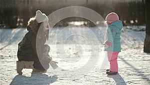 Laughing Woman in Warm Clothing is Throwing Snow at Her Daughter Wearing Snowsuit. Mother and Child Enjoying Cold Sunny
