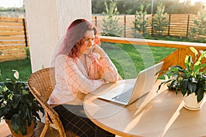 Laughing woman wearing pajama having video call on laptop during lunch break sitting at terrace in her home - summer