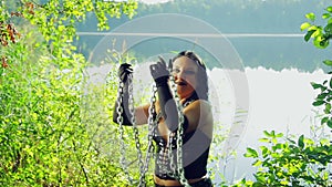 A laughing woman of a witch in black clothes on the shore of the lake is having fun and dancing with chains in her hands