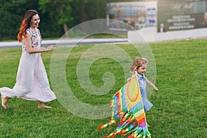 Laughing woman in light dress and little cute child baby girl running, catching up and play with colorful kite in park