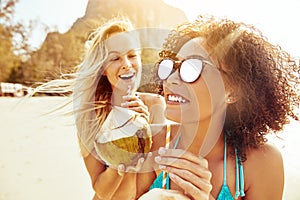 Laughing women walking together along a beach drinking from coco