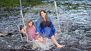 a woman and a young girl are swinging across a fast-flowing river, laughing and splashing with water