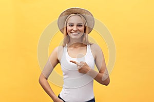Laughing young beautiful woman in t-shirt looking and pointing away with finger over yellow background